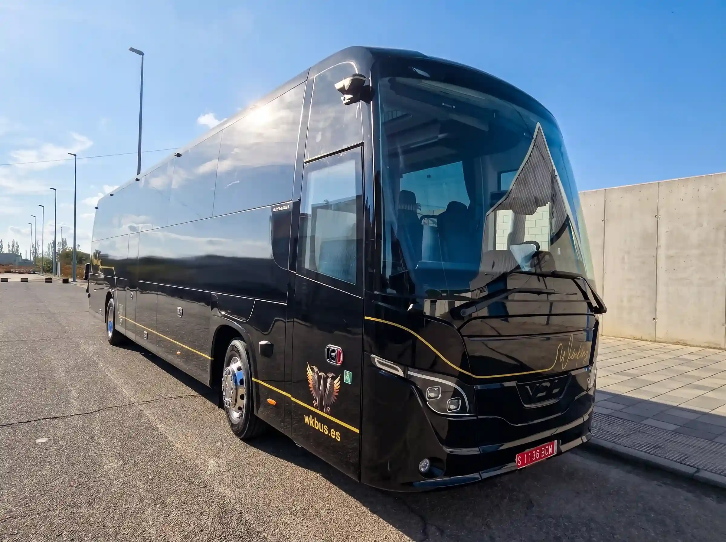 Autocar VDL negro de 55 plazas de Winking Bus visto de frente y de lado, con el rótulo dorado Winking en el frontal y la línea dorada del costado, aparcado junto a un muro de hormigón bajo un cielo azul