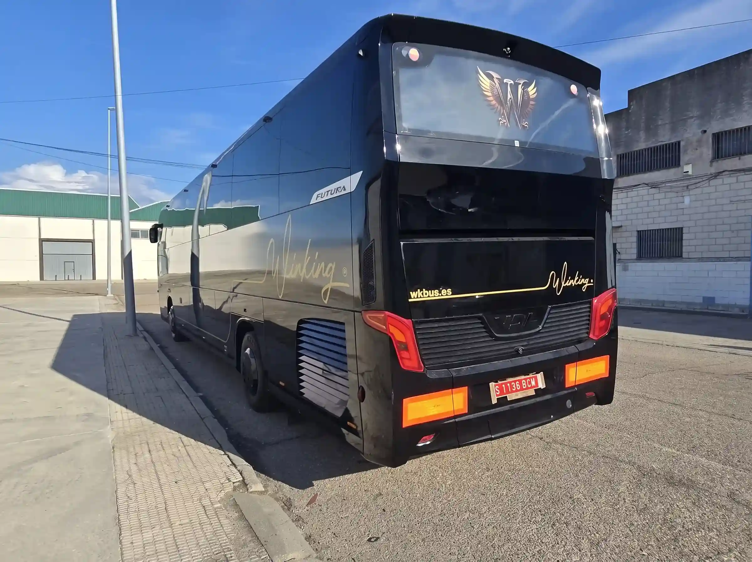 Parte trasera del autocar VDL negro de 55 plazas de Winking Bus, con el escudo dorado de la luneta, los pilotos rojos y naranjas y el rótulo Winking junto a wkbus.es, aparcado ante una nave industrial