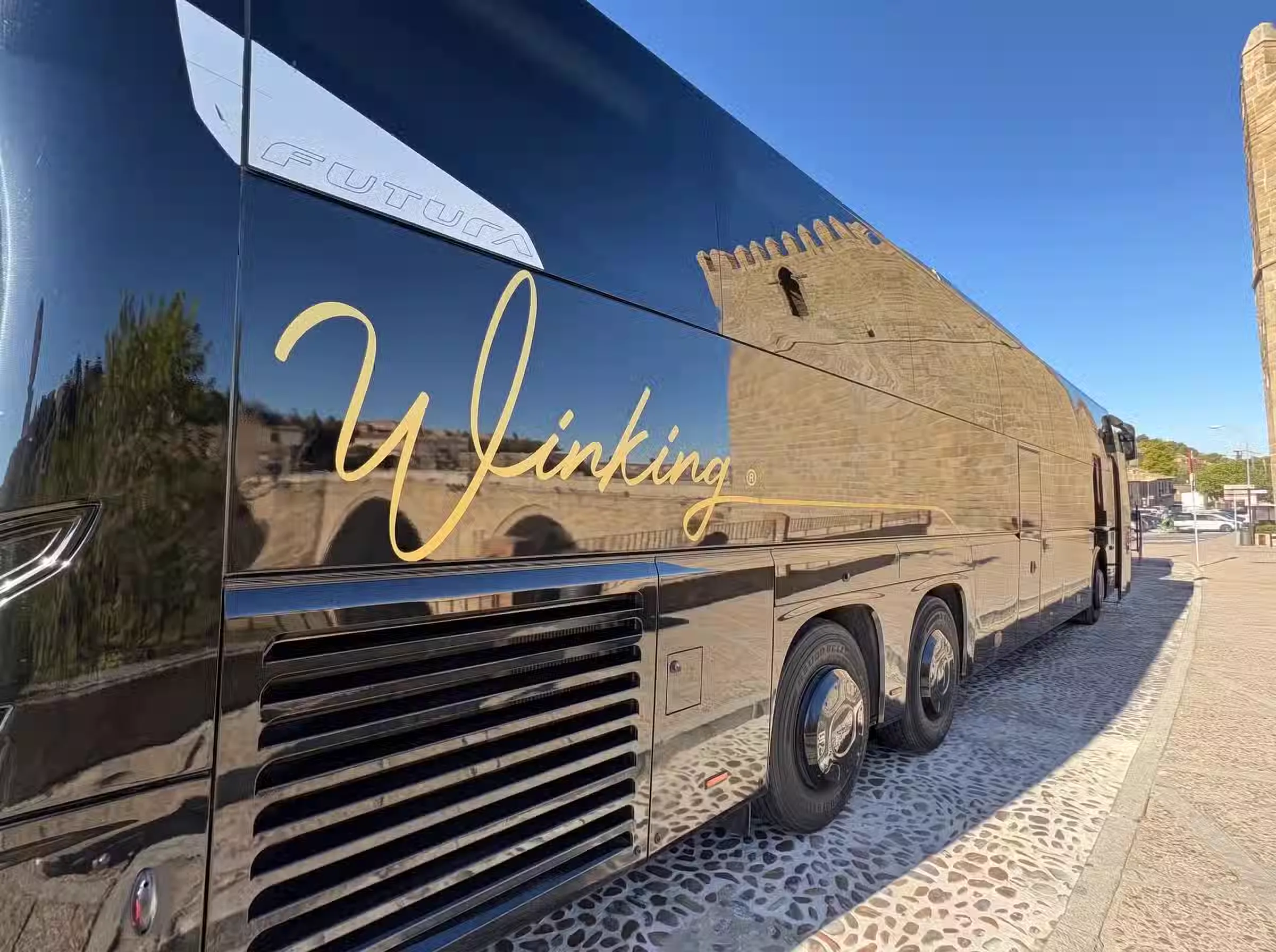 Costado de un autocar VDL Futura negro de Winking Bus con el rótulo dorado Winking, con reflejos de un edificio de piedra y el cielo azul