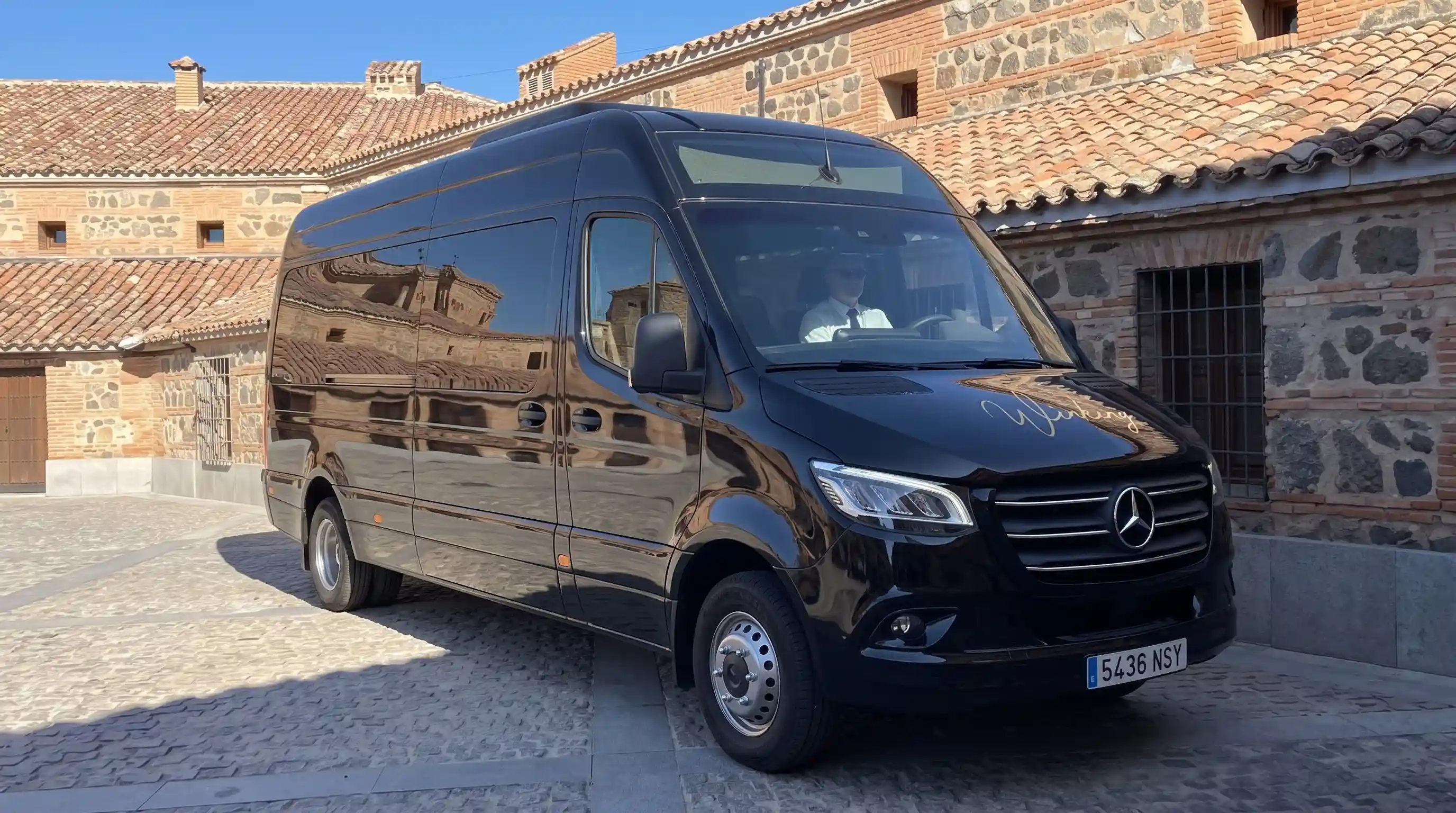 Microbús Mercedes-Benz Sprinter negro de 19 plazas de Winking Bus aparcado en una plaza empedrada de Toledo, de tres cuartos delantero, con el rótulo dorado Winking en el capó y el conductor al volante